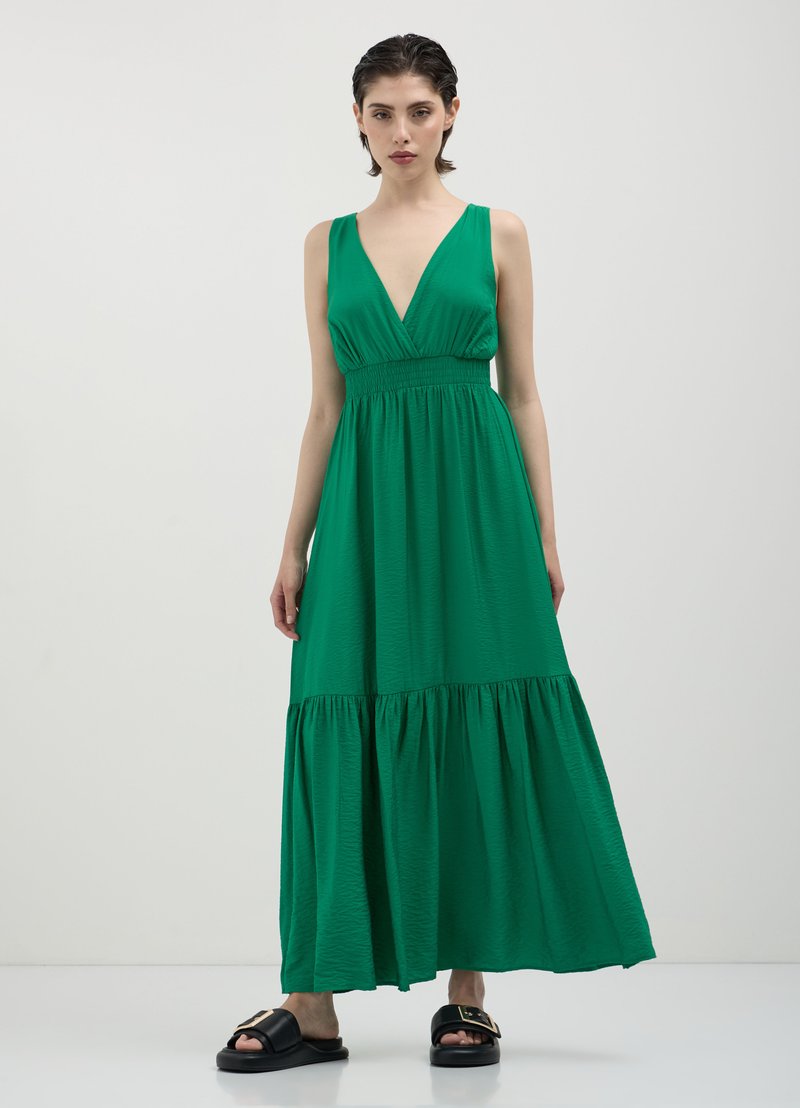 Grünes Maxikleid mit V-Ausschnitt, elastischem Taillenbund und gestuftem Rock. Leichter Stoff mit glatter Textur, der eine fließende Silhouette bietet. Schwarze Sandalen.