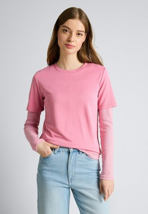 Jeune femme aux longs cheveux bruns portant des tops roses superposés et un jean bleu clair, debout avec une main dans la poche devant un fond uni.