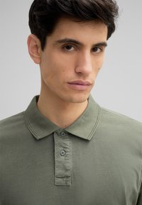 Groene katoenen polo met kraag, voorzien van een knopenlijst met twee knopen en een zachte textuur. Close-up van de halslijn en stofdetails.