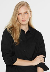 Black button-up shirt in lightweight fabric, featuring a pocket, folded sleeves, and a classic collar, creating a casual yet structured look.