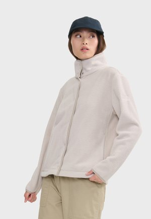 Veste en polaire beige clair avec un col haut, fermeture à pression à l'avant et finitions contrastantes ; associée à une casquette bleu foncé.