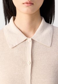 Femme aux cheveux lisses et foncés porte un haut en maille beige boutonné avec un col pointu. Seuls le bas de son visage, son cou et le haut de sa poitrine sont visibles.