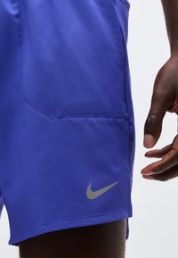 Κοντινό πλάνο ενός ατόμου που φοράει μπλε σορτς Nike με αντανακλαστικό λογότυπο Swoosh, δείχνοντας την πλαϊνή τσέπη και μέρος του χεριού του.