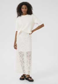Haut blanc à coupe décontractée associé à une longue jupe en dentelle blanche ornée d'un motif floral et des sandales noires à semelles épaisses. Tissu lisse et dentelle texturée.