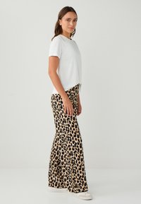 Witte T-shirt met korte mouwen, gecombineerd met tankleurige flared broek met luipaardprint; model draagt witte sneakers en staat tegen een effen achtergrond.