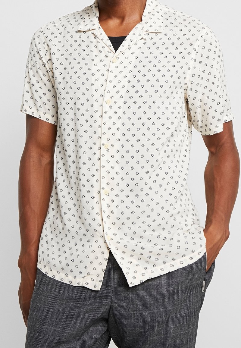 Homme portant une chemise crème à manches courtes boutonnée, ornée de petits motifs cachemire noirs, et un pantalon à carreaux gris, une main dans la poche.