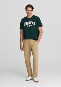 Maglietta verde scuro con la scritta "GREENWICH New York" in bianco, abbinata a pantaloni beige e sneakers bianche, in piedi contro uno sfondo neutro.