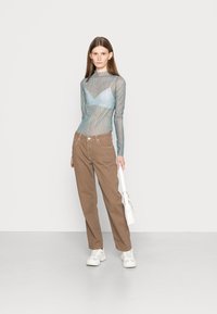 Blusa de manga longa em tom azul claro e tecido transparente com padrão de ondas, combinada com calças retas castanhas e ténis brancos. O modelo segura uma bolsa branca.