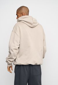 Sudadera de color beige claro con capucha ajustable, puños de canalé y un corte relajado, combinada con pantalones gris oscuro que cuentan con un bolsillo lateral.
