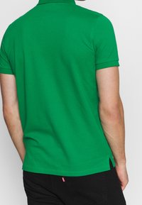 Polo shirt in cotone verde con colletto a costine, maniche corte e spacchi laterali all'orlo, che presenta una texture liscia e di colore uniforme.