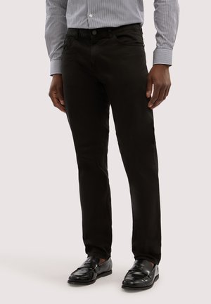 Mężczyzna ubrany w czarne, dopasowane spodnie, czarne skórzane lordsy oraz włożoną w spodnie pasiastą, długorękawową koszulę, stojący na jednolitym tle.