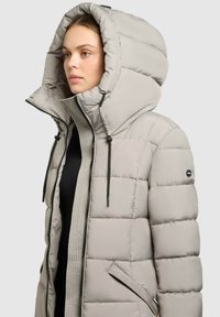 Ljusgrå puffjacka med en stor huva, med dragkedja framtill, två sidofickor och en texturerad innerväv. Förstärkta ärmslut och justerbara dragskor.