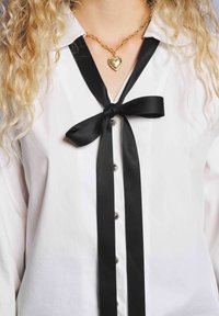 Γυναίκα που φοράει λευκό πουκάμισο με διακοσμητική μαύρη κορδέλα και χρυσό κολιέ με καρδιά, με σπαστά ξανθά μαλλιά.