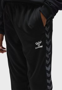 Person trägt schwarze Hummel-Hose mit reflektierenden Chevron-Streifen und hat die rechte Hand in der Tasche vor einem hellgrauen Hintergrund.