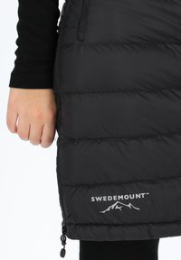 Svart isolerad jacka med quiltat mönster, silverfärgad "SWEDEMOUNT"-logotyp, sidoficka med dragkedja och hög krage.