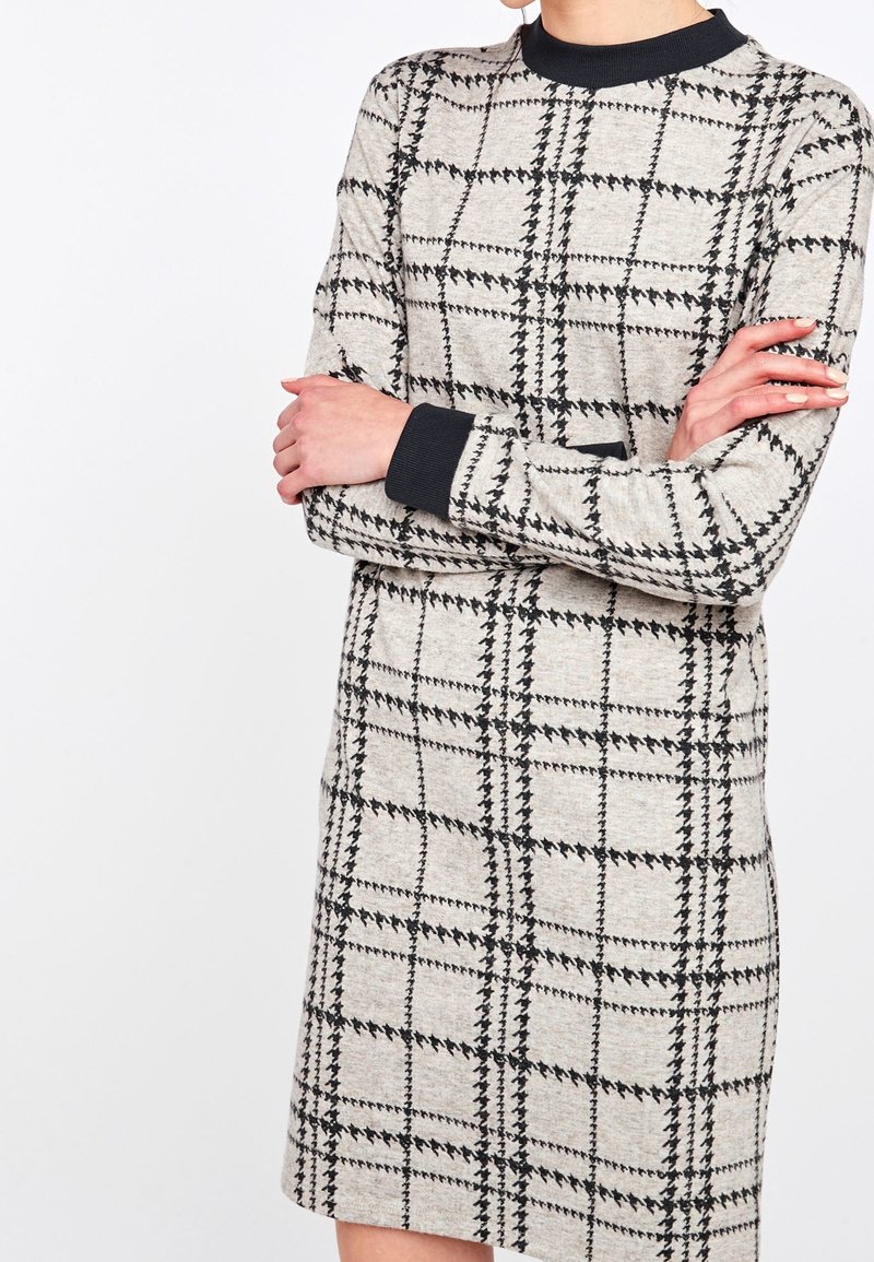 Robe beige avec un motif à carreaux noirs, manches longues et col rond. Le tissu semble doux avec des accents côtelés aux poignets.