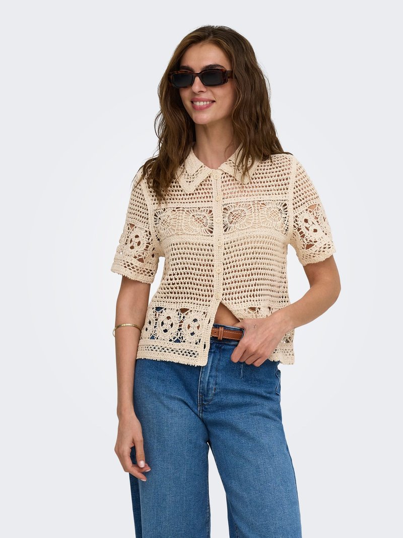 Femme portant des lunettes de soleil foncées, une chemise beige courte à manches courtes en crochet avec des boutons, un jean bleu et une ceinture marron, souriante avec la main près de la taille.