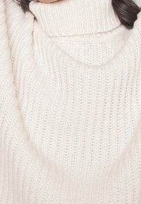 Brede gebreide coltrui in een zachte, gebroken witte kleur. Voorzien van een geribbelde textuur met een losse pasvorm en een brede, gedrapeerde halslijn.