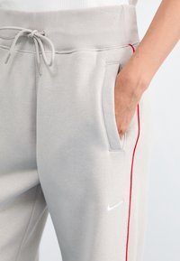 Högra handen i fickan på ljusgrå Nike sweatpants med röd sidostrand och snodd i midjan, på en person som bär en vit topp.