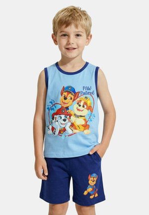 Jeune garçon portant un débardeur bleu Paw Patrol et un short marine, souriant avec une main dans la poche, sur un fond clair uni.