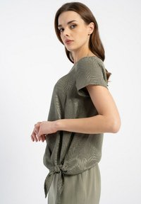 Olive zielona koszulka z krótkim rękawem, z węzłem wywiązanym w talii i subtelnym wzorem liści na powierzchni materiału.