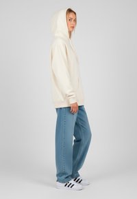 Felpa con cappuccio color crema, con tasca frontale, abbinata a jeans a gamba larga blu chiaro e sneakers in bianco e nero. Tessuto dal finish liscio.