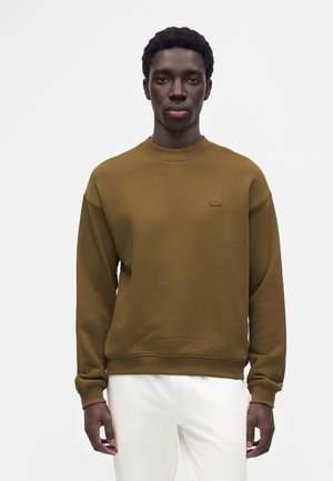 Jeune homme portant un sweat-shirt marron avec un petit logo sur la poitrine et un pantalon blanc, debout devant un fond clair uni.