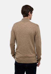 Homme portant un pull léger marron clair à manches longues, de dos, devant un fond blanc uni.