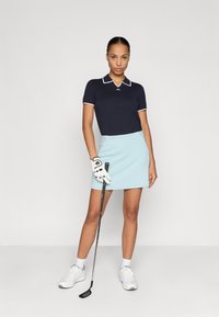 Námorně modré polo tričko s bílým límcem a logem, spárované se světle modrou páskovanou sukní. Hráč drží golfovou hůl, oblečený v bílých sportovních botách a rukavici.