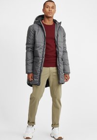 Grå hettegenser-puffjakke med kviltet design, to frontlommer, brukt over en bordeaux strikkegenser, sammen med khaki bukser og hvite sneakers.