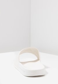 Valkoinen sandaali, jossa on sileä, pehmeä pinta, ympyrän muotoinen avoin takapää ja tasainen pohja. Minimalistinen muotoilu ilman lisäkoristuksia.