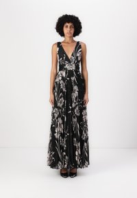 Robe maxi noire à fleurs avec un décolleté en V profond, un tissu plissé et une taille cintrée. Accessoirisée avec de légers motifs floraux et des talons noirs élégants.