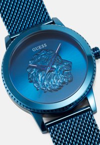 Reloj de metal azul con una correa de malla texturizada, un cabezón de león en relieve en la esfera y un diseño minimalista con elegantes manecillas de horas y minutos.