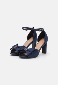 Navy blue heeled sandals with an open toe, satin material, large bow accent on the front, and a buckle strap around the ankle.