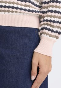 Adīts džemperis ar ģeometrisku rakstu rozā, tumši zilā un olīvu toņos, ar ribas aprocēm un mīkstu tekstūru. Zem tā zils džinsu audums.