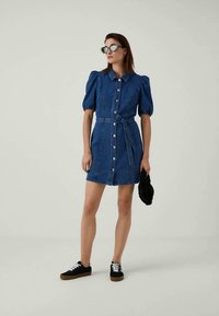 Mujer vestida con un vestido de mezclilla azul de botón frontal, con mangas abullonadas y cinturón, zapatillas negras, gafas de sol negras y sosteniendo un accesorio negro de peluche.