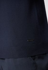 Dunkelblaues Sweatshirt mit feiner Strickstruktur, geripptem Saum und einem kleinen metallischen Markenetikett am unteren Rand. Kombination mit dunklen Hosen.