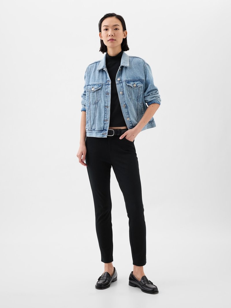 Femme portant une veste en denim bleu clair sur un col roulé noir, un pantalon noir avec une ceinture, et des mocassins noirs, se tenant devant un fond blanc uni.