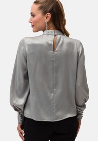 Blusa argentata a maniche lunghe con collo alto, dettaglio a fessura sulla schiena e rifiniture con bottoni ai polsini. Tessuto liscio e lucido.