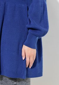 Blå stickad tröja med ribbstruktur, långa ärmar och sidofickor. Den nedre kanten är avslutad med en mjuk ribb. Handen vilar på tyget.