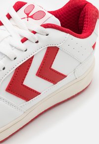 Sneakers bianchi con accenti rossi, caratterizzati da una tomaia texturizzata, un design a zigzag a contrasto e un colletto imbottito. Dettagli cuciti e logo presente.