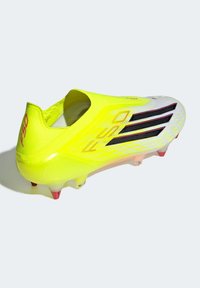 Φωτεινό κίτρινο ποδοσφαιρικό παπούτσι με υφή στην επάνω επιφάνεια, μαύρες ρίγες και διαφανές πέλμα με κόκκινες τάπες. Λεπτός, ελαφρύς σχεδιασμός.