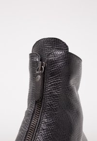 Detailní záběr na černou koženou botu s texturou, vertikálním zipem a poutkem na tažení na jednoduchém bílém pozadí.