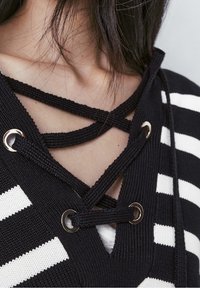 Top rayé noir et blanc à encolure lacée avec œillets en métal et cordons tressés, mettant en valeur un tissu tricoté texturé.