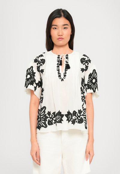 Jonge vrouw in een witte blouse met korte mouwen met zwarte bloemenborduursels en bijpassende witte shorts, staande tegen een effen achtergrond.