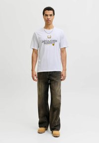 Camiseta de algodón blanca con texto gráfico en negro y amarillo, combinada con jeans rectos de mezclilla oscura deslavada y calzado beige. Collar de cadena de plata.