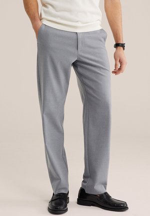 Moški v svetlo sivih hlačah po meri, črnih usnjenih mokasinih, belem pletenem puloverju in črni ročni uri, stoji z rokami delno v žepih.