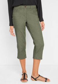Pantalon court vert olive en mélange de coton, doté de poches avant, d'une fermeture à bouton et d'une texture lisse. Porté avec des sandales noires.
