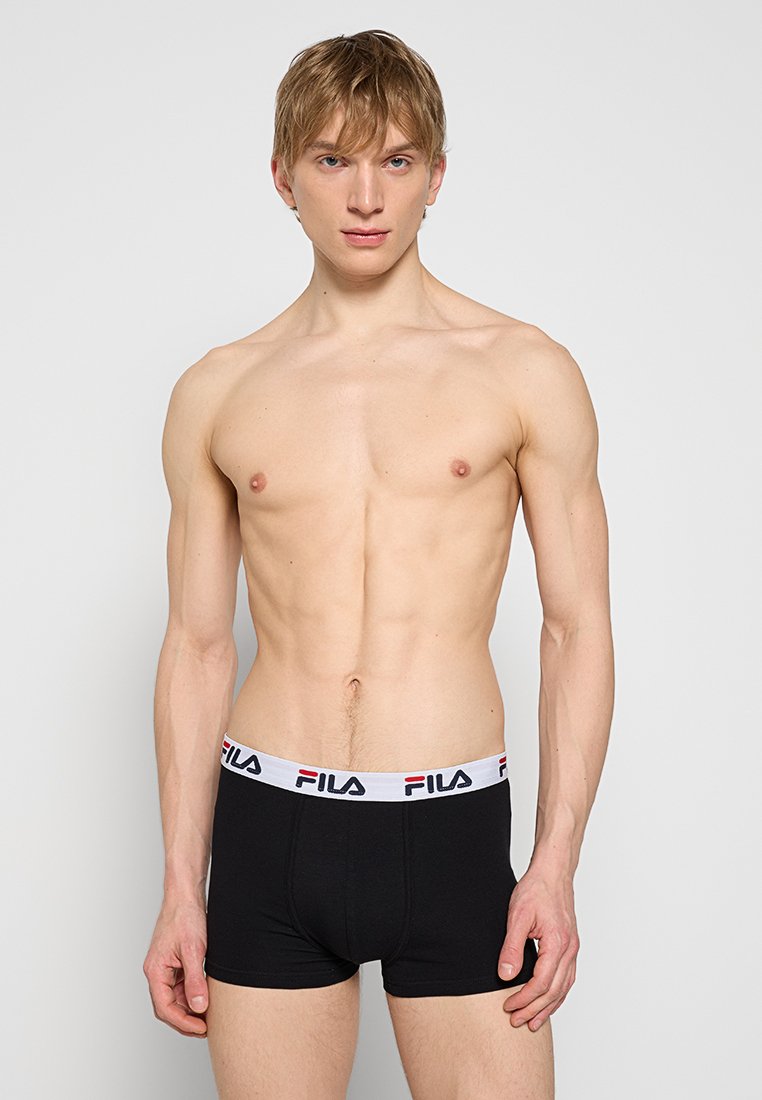 Jeune homme debout portant un boxer noir Fila avec une ceinture blanche siglée sur un fond clair uni.
