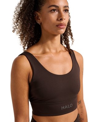 Ung kvinde med krøllet hår iført en mørkebrun sports-bh med "HALO"-logo, vendt mod højre mod en hvid baggrund.
